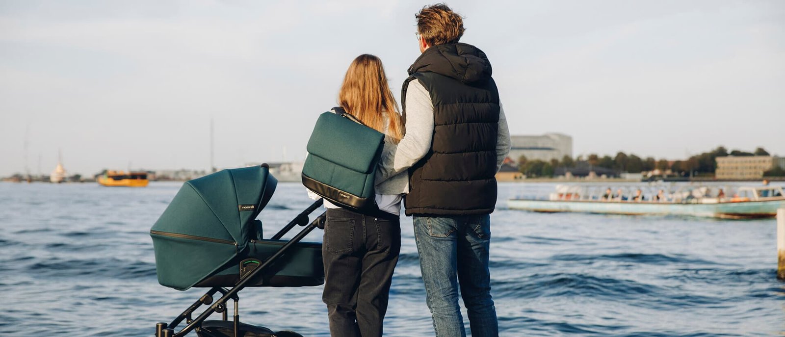 Couple with a green pram looking out over the sea from a waterfront, one parent wearing a backpack
