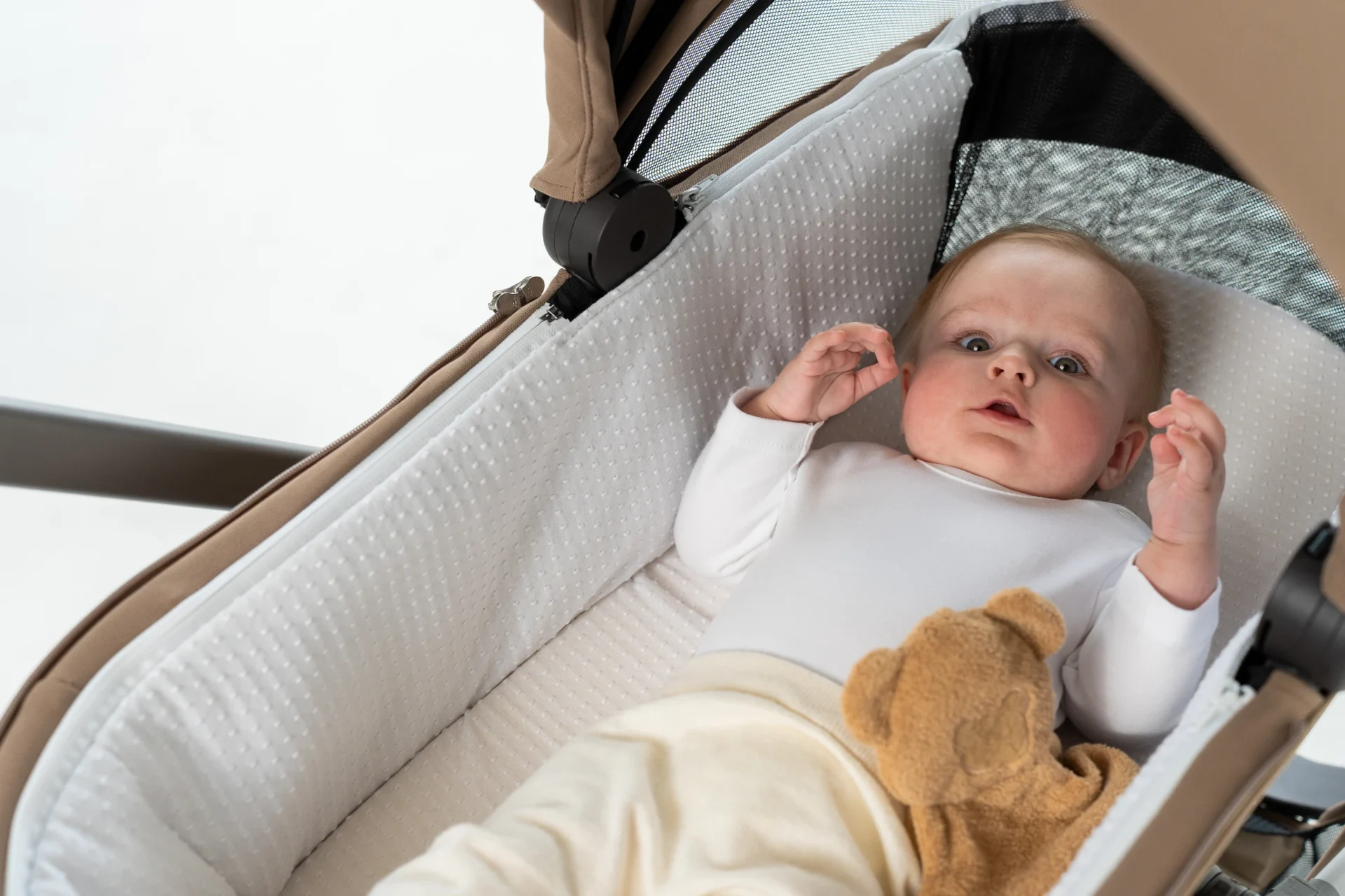 Baby lying in a soft carrycot looking up, a small teddy bear beside them