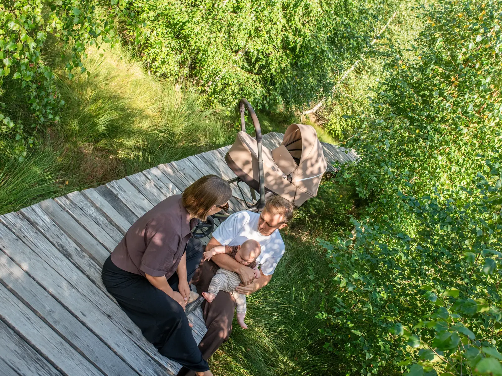 Family resting on a wooden boardwalk in lush green woodland, looking down at their baby, pram behind them