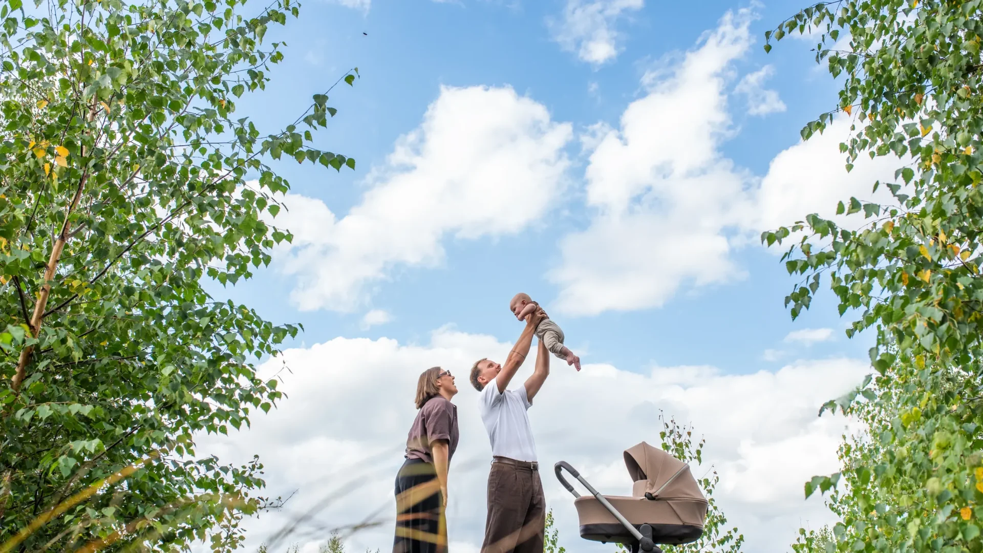 Parents lifting their baby up into a bright blue sky, framed by birch leaves